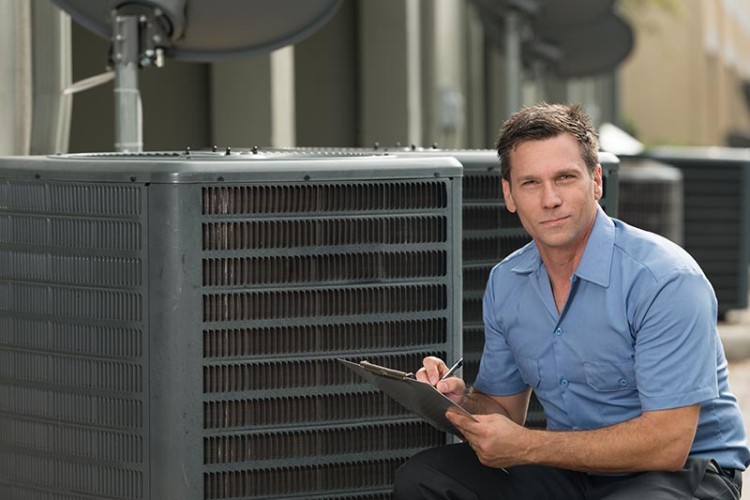 AC Technician kneeling next to an AC unit with a clipboard in his hand.